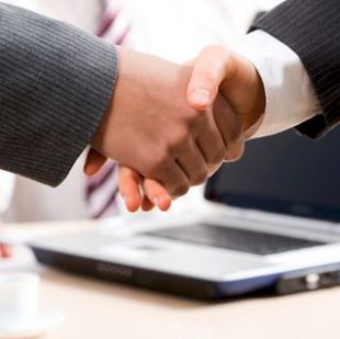 Zwei Geschäftspartner besiegeln ihre Zusammenarbeit mit einem Handschlag, während im Hintergrund ein Laptop steht.