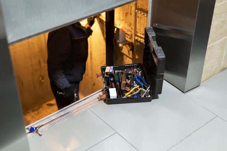In der unteren Hälfte des Bildes sieht man einen Monteur der in dem beleuchteten Aufzugsschacht steht und an der Kabineninstallation arbeitet. Ein Teil der Aufzugskabine ist in oberen Teil des Bildes über dem Monteur zu sehen. 